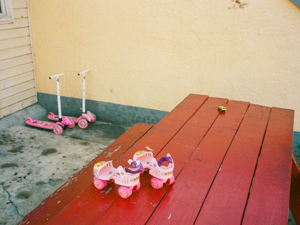 Photograph by Casey Joiner of red picnic table with a pair of pink children's roller blades in front a light yellow wall in a yard/ porch setting. The ground is a stained green concrete and two pink children's scooters with white handles sit in the left corner of the image.