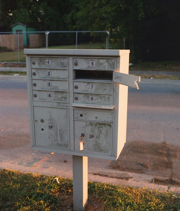 Photograph by Casey Joiner of a large white mailbox with eleven comaprtments in front of the street around sun down. The street is painted by the warm pink/ orange hues of the sun.