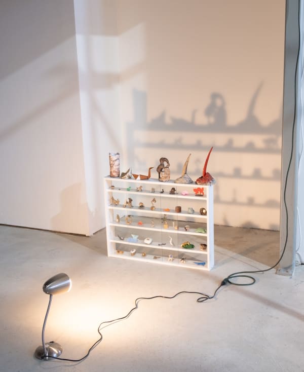 A sculpture by Jon Gott composed of a small shelf with many made and found tchotchkes organized throughout. A metal desk lamp shines directly at the assemblage, casting a dramatic shadow behind.