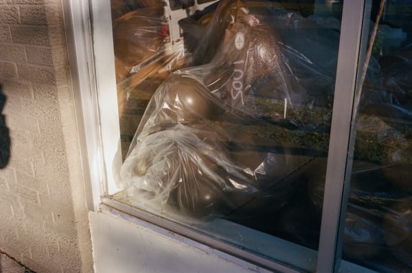 Photograph by Casey Joiner of a window revealing clear plastic bags full of black balloons. The reflection of the glass overlays the image of a trash bin.