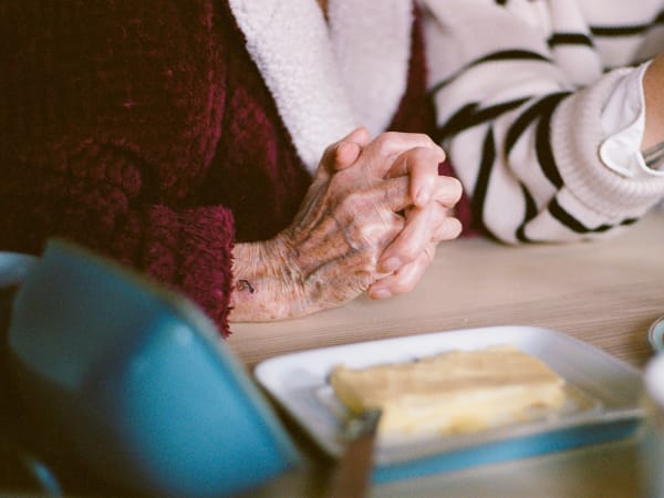 Photograph by Casey Joiner of an elderly person in a red robe's hands with interlocked fingers rested on the table in front of a stick of table butter and the end of a knife. There is another person frame but their hand and body are cut off by the frame.