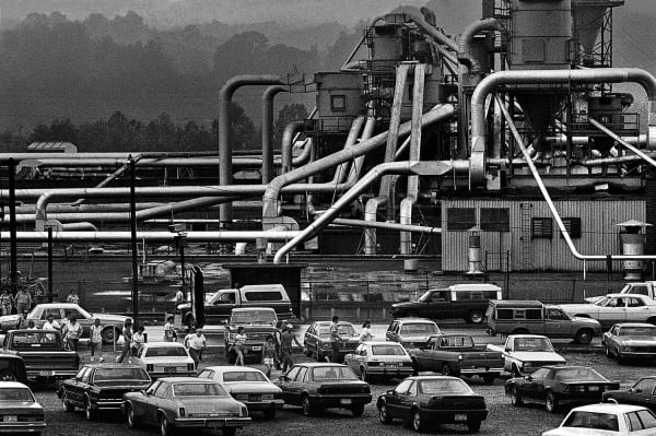 A black and white photograph by Spencer Ainsley shows the Bernhardt Furniture factory workers exit a plant on a rainy day in Lenoir, North Carolina. The factory looms behind them.