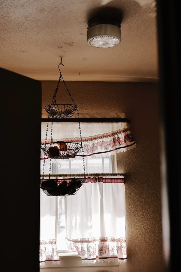 Photograph by Casey Joiner of a kitchen setting with a hanging basket of food stuffs in front of white curtains with light shining through.