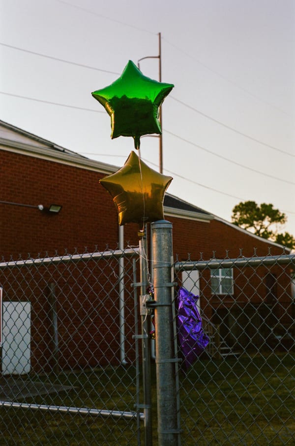 Photograph by Casey Joiner of three star shaped balloons in Mardi Gars colors (Green, Yellow, Purple) tied/ caught on chain link fence in front of a redbrick house at dusk.