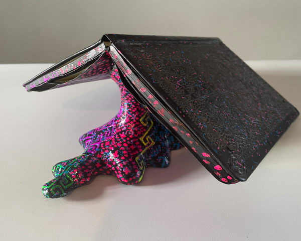 Lasercut design of sugar skulls on the cover of a moleskin that is open and resting on top of a colorful organic shaped sculpture