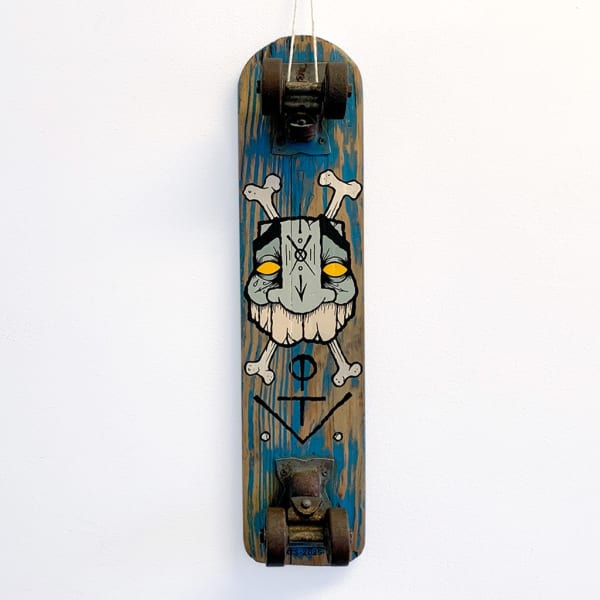 A vintage wood skateboard with a painted image of a masculine face with to bones in an "x" behind the head; replicating a skull and crossbones. Blue paint drips down the board in the foreground.