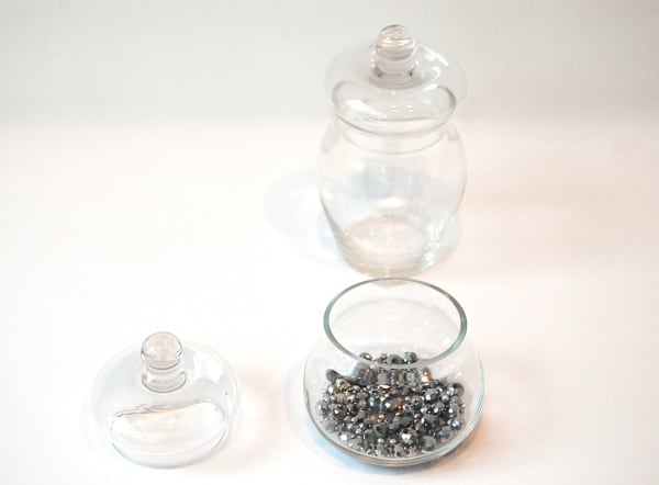 Two glass jars, one empty with lid on and one with many silver beads and lid sitting next to it