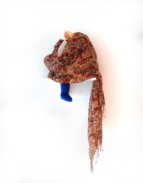 Wall sculpture with patterned red scarf wrapped around a toy with a blue foot sticking out from beneath