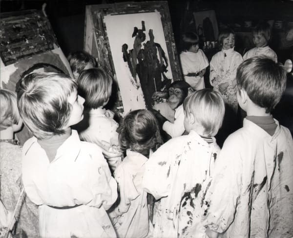 Black and white photograph of children gathered around a chimpanzee painting on an easel