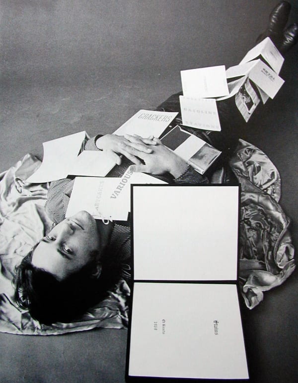 Black and white image of dark haired man lying underneath pile of books