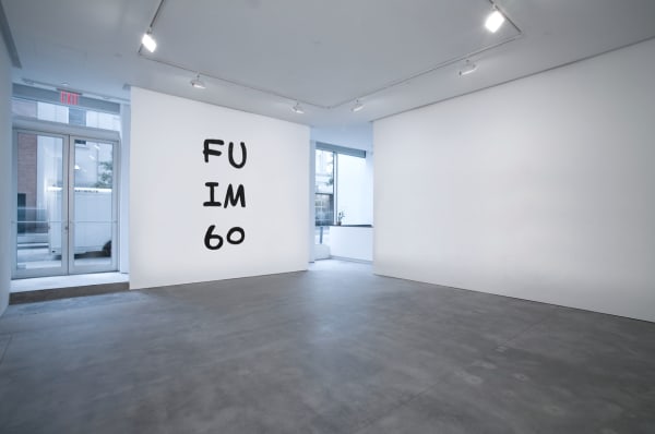 An installation view of a large gallery with a concrete floor and high ceilings containing a wall mural that reads "F U IM 60" in three rows, like an eye chart