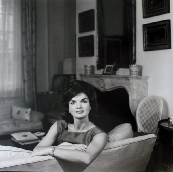 Black and white portrait photograph of Jacqueline Kennedy as she looks over the back of a couch in a domestic interior