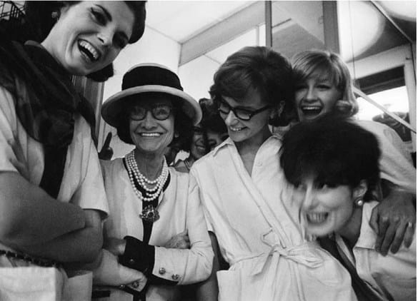 Black and white photograph of designer Coco Chanel, models, and staff gathering around and laughing in the House of Chanel