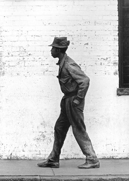 Black and white photograph of a man in profile walking in front of a white brick background