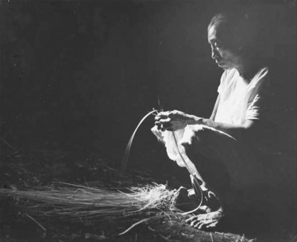 Black and white photograph of a dramatically illuminated Mayan person weaving a palm hat in profile in a completely dark setting