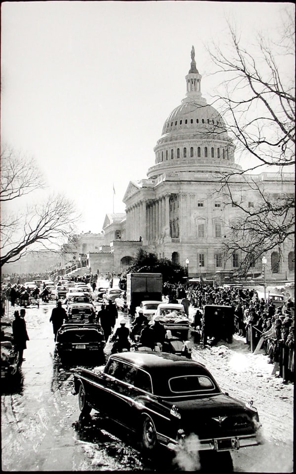 Black and white photograph of cars driving past crowds of people gathered forJohn F Kennedy's inaugural parade by the Capitol building in Washington DC