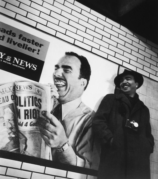 Black and white photograph of Roy Shatt smiling in front of a wall and poster depicting himself holding a newspaper