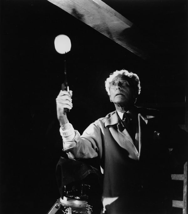 Black and white photograph of Jean Cocteau holding a light in a dark room and looking up to the left