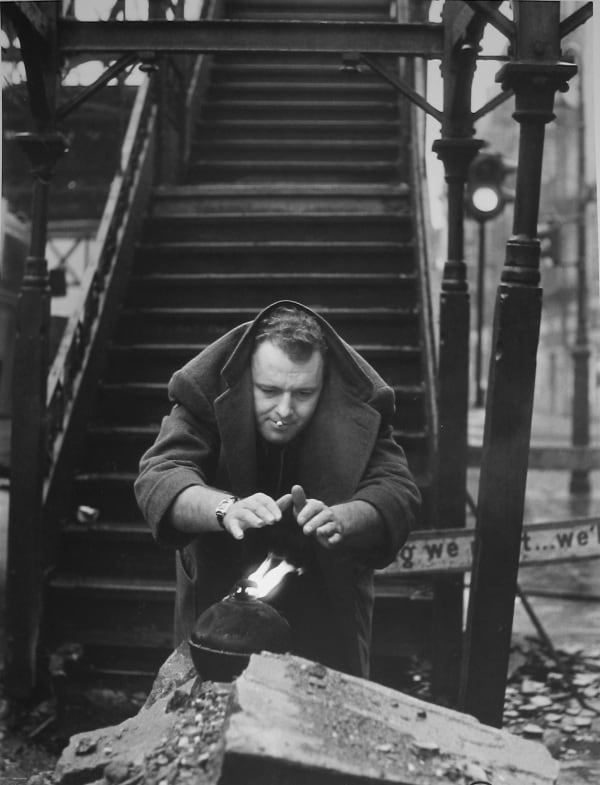 Black and white photograph of Rod Steiger with his jacket over his head, warming his hands over an open flame atop of concrete rubble in front of the steps leading to an elevated train platform in New York City
