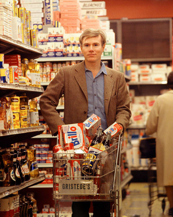 Photograph of Andy Warhol standing in a grocery store with a shopping cart full of items and looking at the viewer