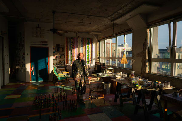 An artist, Steve Silver, standing at a work table amidst paintings and sculptures in an industrial loft setting flooded with late afternoon light