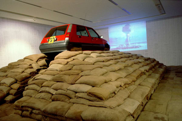 Art installation, a red Renault car on top of a pyramid of sand-filled jute sacks, video projection of atomic explosion