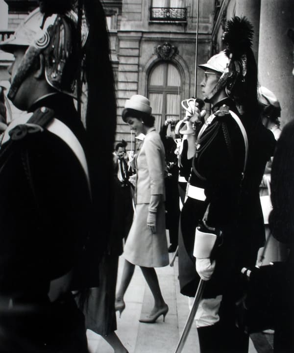 Black and white photograph of Jackie Kennedy in profile walking down steps in Paris surrounded by soldiers