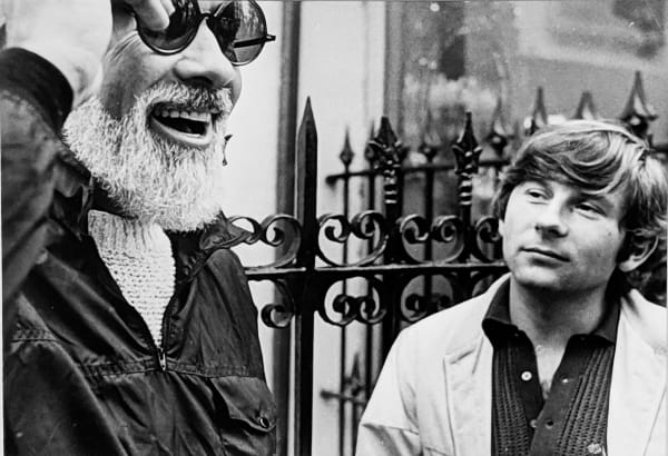 Black and white photograph of William A. Fraker wearing large black round sunglasses and laughing, with Roman Polanski looking at him in the background, New York City, on the set of “Rosemary's Baby”