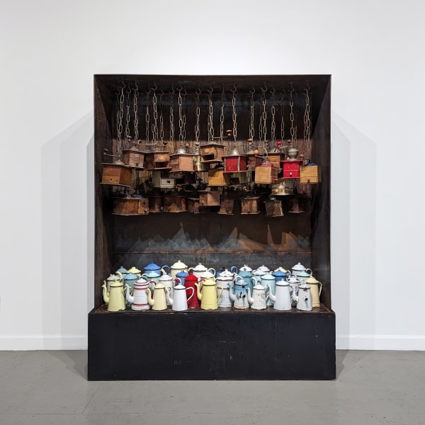 A steel box with an accumulation of wood coffee mills hanging from chains from its top interior and numerous enamel coffee pots at its base