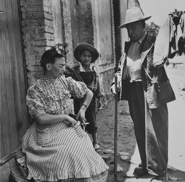 Black and white photograph of artist Frida Kahlo reaching into the pocket of her dress to pay for fabrics being shown to her by a man with a cane and a young boy