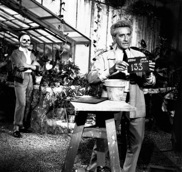 Black and white photograph of Jean Cocteau outside in a garden holding a sign that reads “133” and Edouard Dermithein skull makeup looking at him in the background