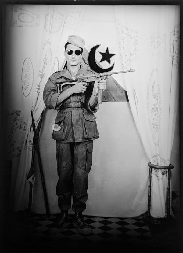 1960s black and white photograph of a man standing holding a gun while wearing sunglasses and a military uniform standing in front of the Algerian flag