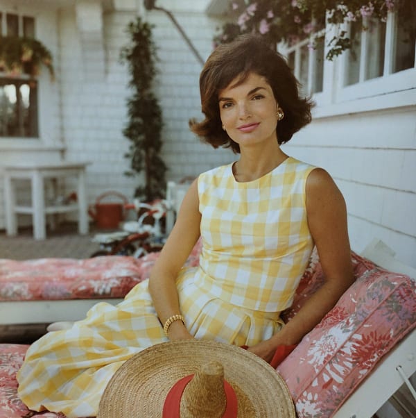 Color photograph of Jackie Kennedy in a yellow gingham dress sitting on a cushioned lawn chair in front of a white house