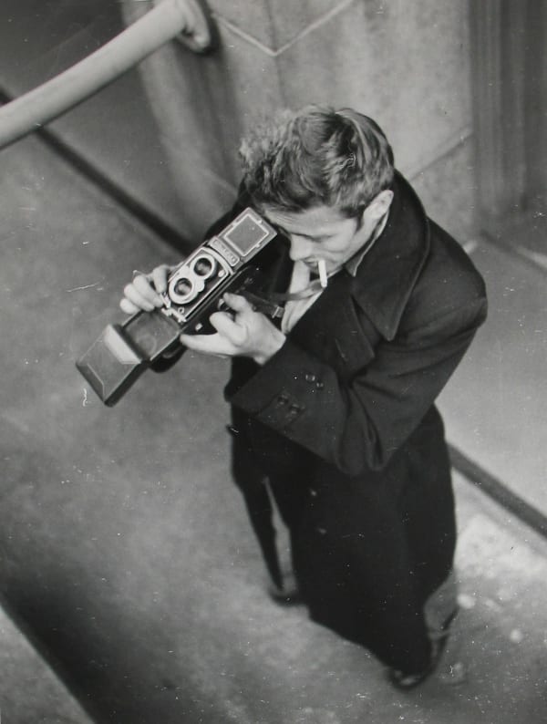 Black and white photograph of James Dean taken from a higher vantage point as Dean is aiming his Rolleiflex camera upwards on a New York City street