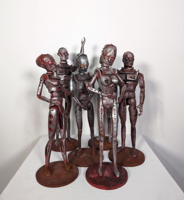 A group of six rust and silver-colored robot figurines, two with male anatomy and four with female anatomy, stand in varying positions on a white pedestal.