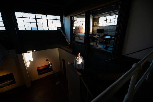 An artist, Curtis Mitchell, hit by a small patch of sunlight as he stands on a staircase in the center of a dark loft setting