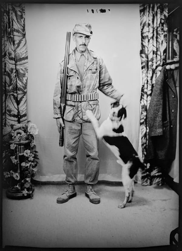 1960s black and white photograph of a soldier standing wearing his army uniform while petting a dog to the right of him that is jumping on its legs