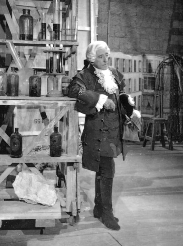 Black and white photograph of Jean Cocteau in Louis XV costume, wearing a white curled wig, ruffled collar, and coat, leaning against a wooden set piece holding a variety of glass bottles