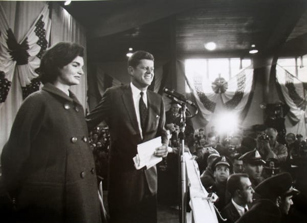 Black and white photograph of John and Jackie Kennedy addressing a crowd from an elevated platform inside a venue