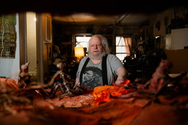An artist, Michael Sullivan, sitting at his work table amidst figurative robotic sculptures in a dimly-lit loft setting