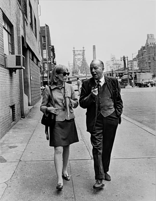 Black and white photograph of Mia Farrow and Maurice Evans taking a break, walking in lower Manhattan with a view of the bridge behind them, New York City, on the set of “Rosemary’s Baby”