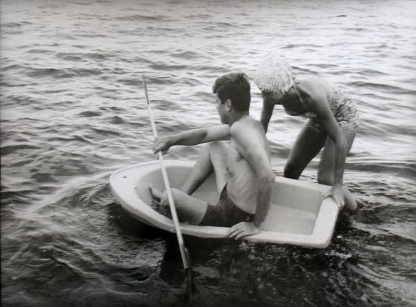 Black and white photograph of Jackie pushing John F Kennedy in a small boat in the water at a Hyannis Port beach