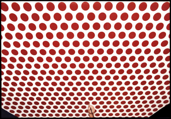 Photograph of a hand with a paintbrush painting a red Ben-Day dot on a canvas filled with identical red dots, arranged equidistantly