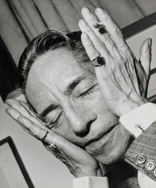 Black and white photograph taken at a diagonal angle of Mexican composer Agustin Lara stretching his face with both hands as he holds a lit cigarette between two fingers