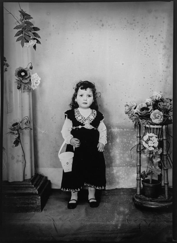 1960s black & white photograph of a young girl wearing an embroidered dress, in a photography studio with artificial flowers