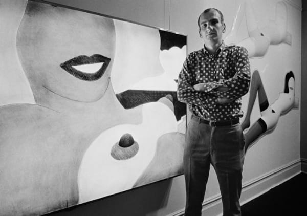 Black and white photograph of Tom Wesselmann standing with folded arms in front of a painting of a female nude