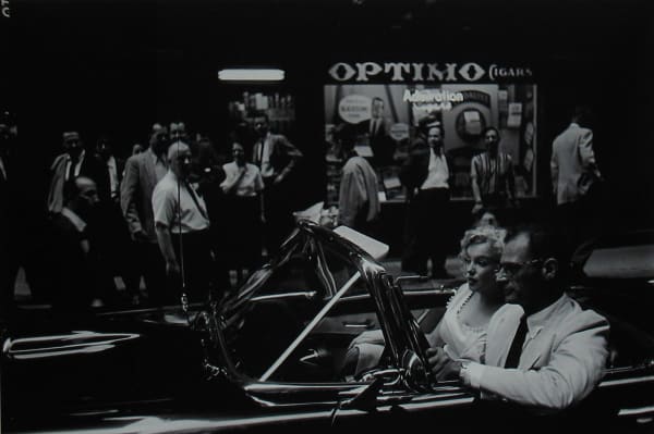 Black and white photograph of Marilyn Monroe and playwright Arthur Miller driving a Thunderbird convertible down a street filled with onlookers