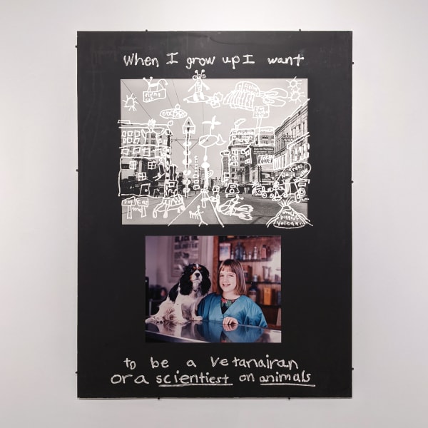 Two photographs on a black board, one is black and white of a street with white child-like doodles on top of it, the other is a young boy holding a book, the words “When I grow up, I want to be a veterinarian or a scientist on animals,” written in silver pen on the top and bottom of the page
