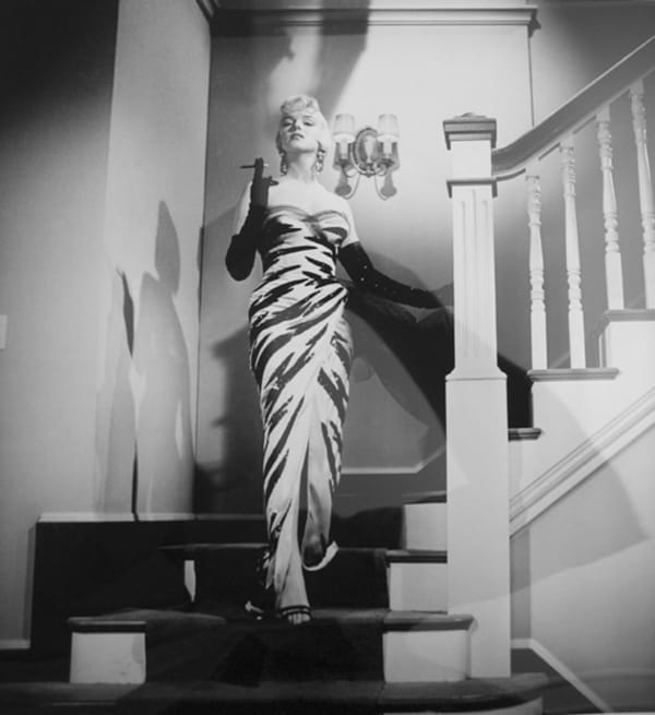 Marilyn Monroe in a white and black animal print dress descends a staircase while smoking a cigarette
