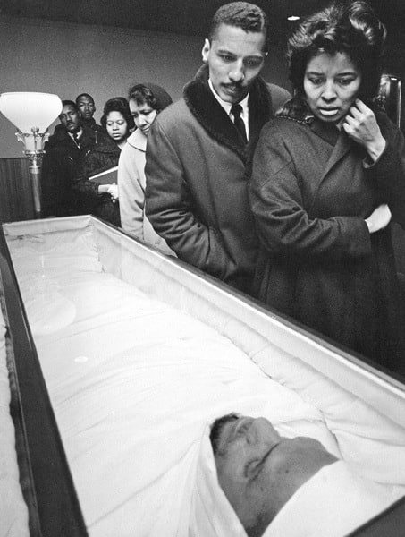Black-and-white photograph of Malcolm X in his casket, shrouded in accordance with Islamic tradition, as African American men and women file by at the funeral viewing